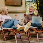 cheerful couple talking in the camper van yard sitting on deck chairs with book and laptop