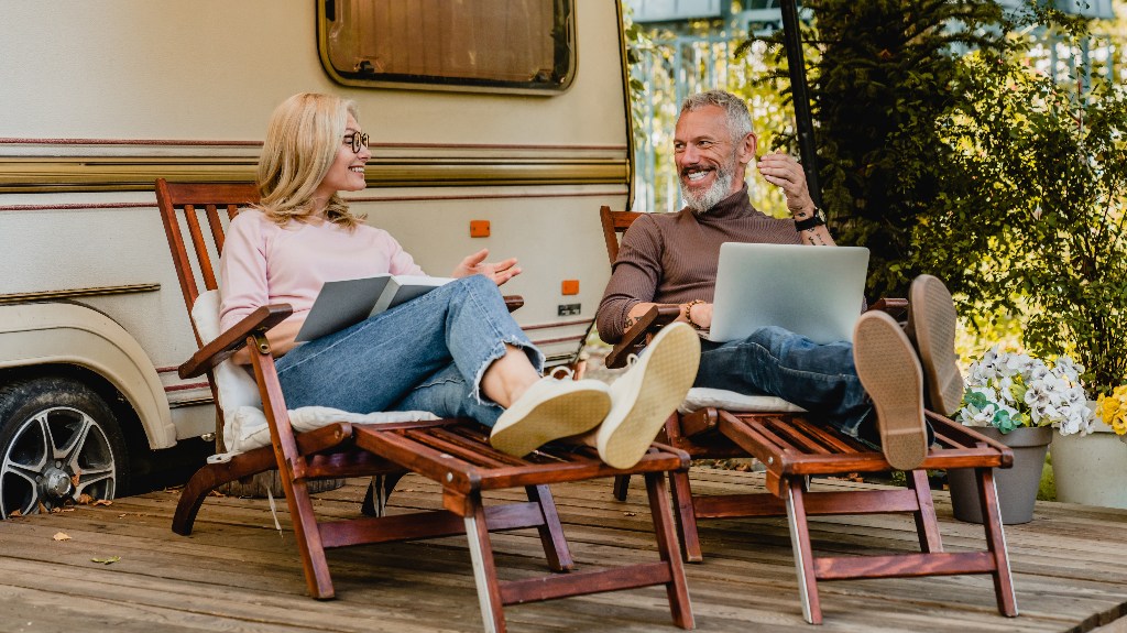 cheerful couple talking in the camper van yard sitting on deck chairs with book and laptop